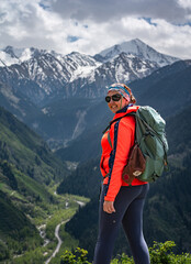 Smiling female hiker in a bright orange jacket and backpack enjoys a stunning view of snow-capped mountains and a lush green valley. Perfect for adventure, travel, and active lifestyle themes © Marina Seagull