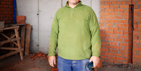A close-up cropped shot of an anonymous construction worker's torso wearing a stained green fleece sweater and blue trousers, holding a work glove against an unfinished brick and concrete wall. © Aigul Sabirova