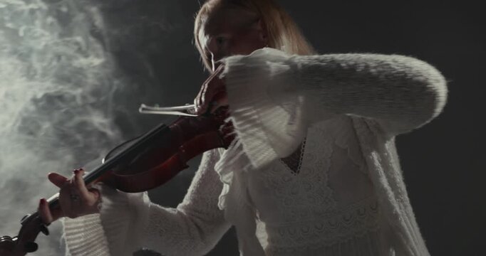 Video frame of a violinist in a white sweater playing in smoky studio light, with soft backlighting, dark background, atmospheric haze, and copy space