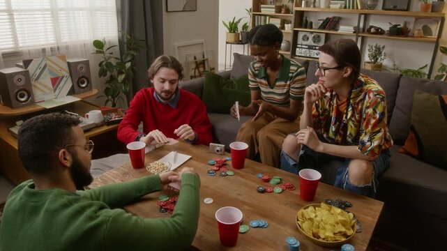High angle shot of four friends playing cards, sipping drink, eating snacks and relaxing in cozy living room