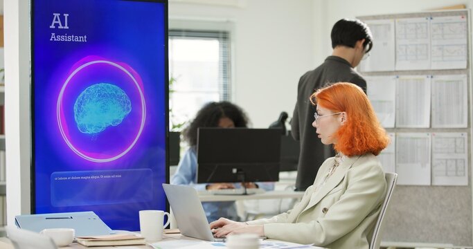 Businesswoman or programmer working on a computer while looking at AI assistant interface on monitor at office. Smart workplace automation, future software engineering, machine learning concept.