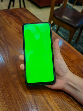 Hand holding blank green screen smartphone screen over warm wooden table. A perspective shows a person holding a mobile phone with a glowing, empty green scren display against a polished wood surface.