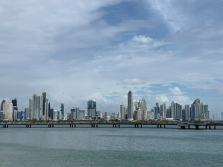 Naklejka premium A scenic view of the Panama City coastal skyline stretching across the bay, viewed from across the water with a long bridge in the foreground against a bright, cloudy daytime sky.