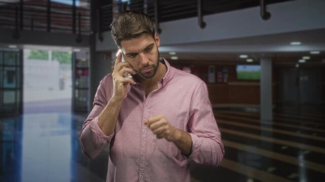Hispanic man with beard in pink shirt holds smartphone to ear, points finger and tilts head in airport; urgent concern.