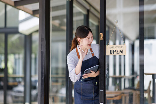 Asian female cafe owner smiling and greeting customers at the open shop entrance, holding a tablet beside the welcome sign, conveying friendly hospitality and small business success