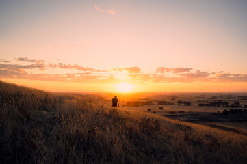 A lone figure surrounded by tall golden grass, stands on a sunlit hillside watching the sun setting above the expansive farm land near the horizon, Mount Noorat, Victoria, Australia © Quincy