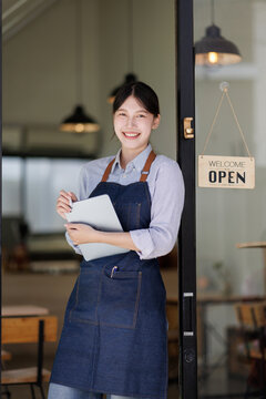 Young asian woman small business owner smiling and holding a tablet, standing proudly at the open entrance of her cafe, ready to welcome customers and manage operations