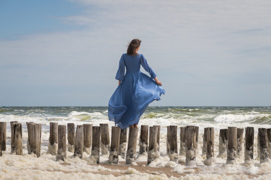 artistic portrait of woman in blue dress standing on wooden groynes on beach at sea