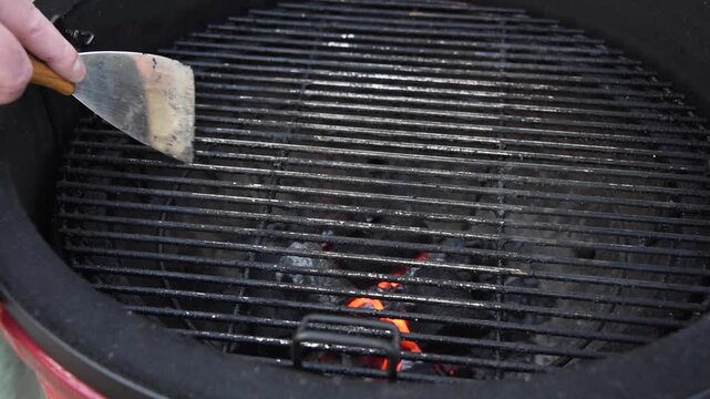 Person uses a metal spatula to clean and scrape the hot grate of a charcoal barbecue grill, preparing it for cooking outdoors