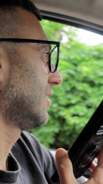 Vertical: A young driver holds his smartphone to record an audio message while behind the wheel, a reckless and illegal behavior that dramatically increases the risk of a road accident.