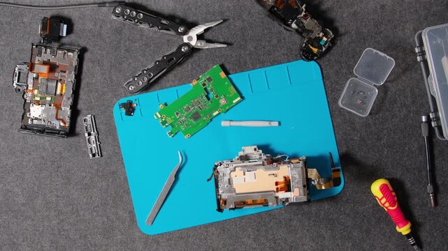 Disassembled camera body and circuit board with various specialized tools for electronics repair and maintenance on a gray mat.