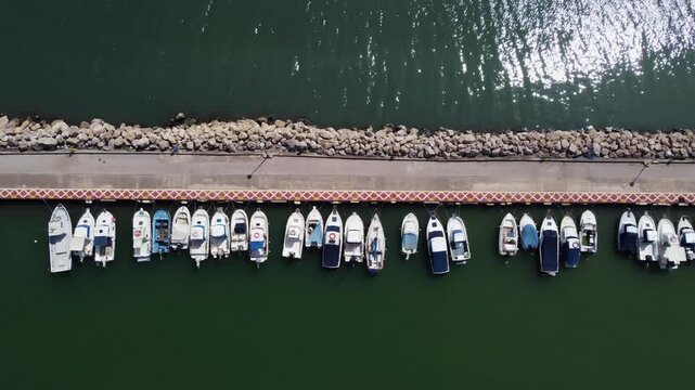 4K top-down aerial video tracking laterally over a stone breakwater and pier. The nadir shot captures a perfectly aligned row of small white boats moored in the calm dark green water.