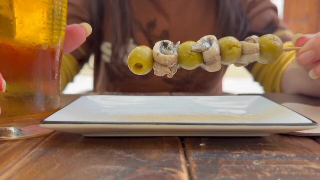 Spanish tapas: Closeup view of the upper body part of a caucasian young woman sitting in a terrace eating anchovies and green olive skewer and drinking a beer. Low angle view.