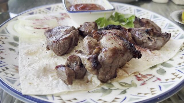 A close-up shot of a plate of shashlik, grilled meat skewers, served with lavash bread, red onion, sumac, tomato sauce, and fresh herbs