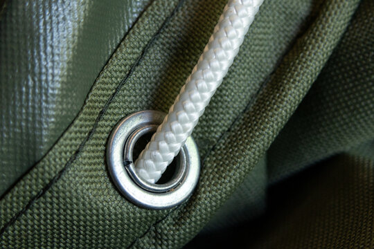 Macro shot showing green heavy duty canvas tarpaulin fabric texture details metal eyelet ring white nylon rope threaded through secure tie down equipment outdoors reliable strong steady connection