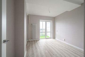 Obraz premium A bright empty room with light grey walls, light wood laminate flooring, white ceiling, a white radiator, and a large glass door leading to a balcony