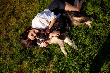 Obraz premium Woman lying on green grass with a dog, embracing and playing together in a sunny outdoor setting, showcasing a joyful bond between pet and owner