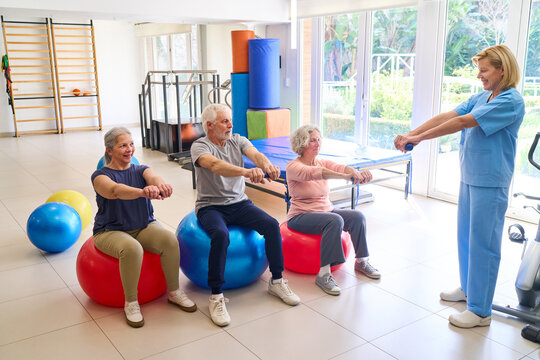 Senior Adults Doing Exercises On Fitness Balls With Physiotherapist In Rehab Center For Recovery And Strength Training Indoors
