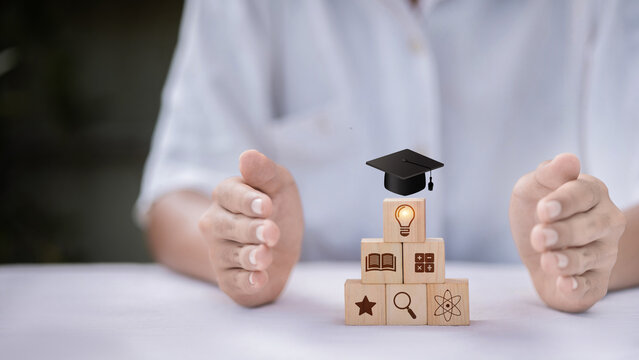 Hands Protecting Pyramid of Wooden Cubes with Education Icons &mdash; Graduation Cap, Lightbulb, Book, Science, and Knowledge Building Concept