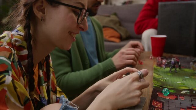 Close up shot of young adult woman in glasses using camera to take picture of her friend, playing tabletop game with group of friends