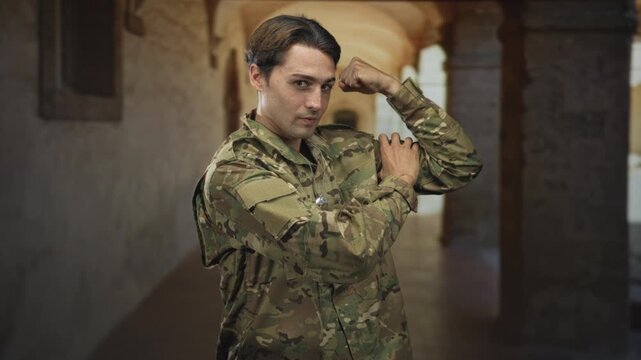 Soldier man in camouflage uniform flexes a bicep and displays dogtags while standing in an arched building corridor with columns; pride duty.