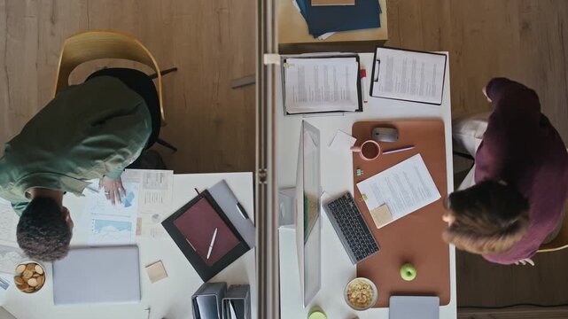 Top view of two male coworkers taking seats at separate office desks and beginning paperwork in modern workspace