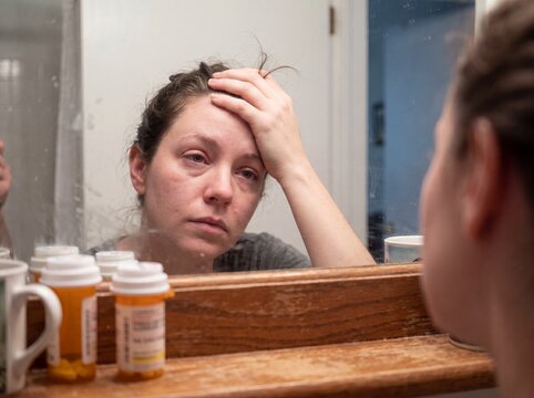 A detail shot of a woman suffering from hypothyroidism reflected in the bathroom mirror early in the morning, her face slightly puffy and eyes tired as she gathers her hair, with several broken strand