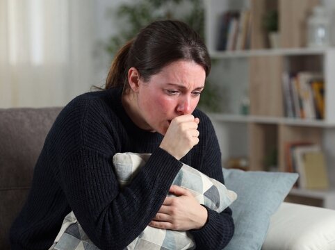 A medium shot of a woman caught in a severe coughing fit due to acute bronchitis, her torso leaning forward with a hand over her mouth and her face flushed from the exertion while clutching a pillow,
