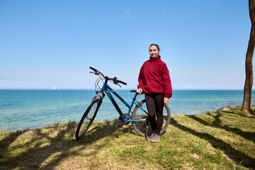 Fototapeta premium Young woman standing with blue bicycle on grassy coast under clear blue sky