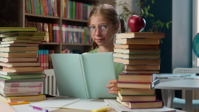Motivated cute Caucasian little girl child school kid in glasses reading book looking at camera daughter elementary primary learning studying in library pupil reader read study learn holding textbook