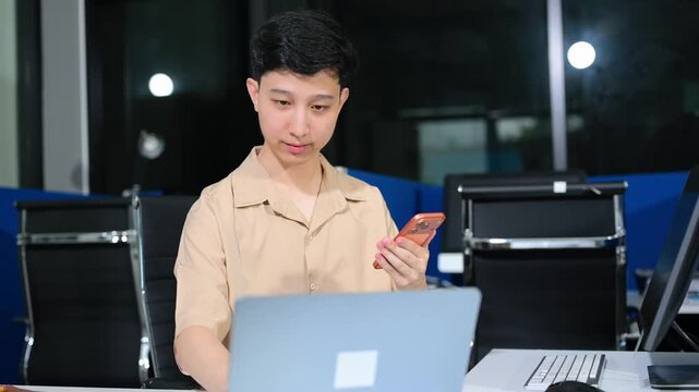 Young Asian businessman using a smartphone app for digital verification while working on a laptop late