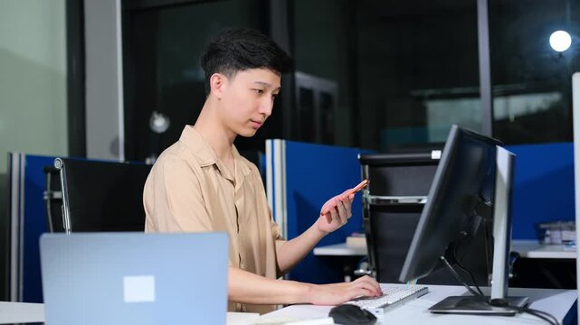 Young Asian businessman using a smartphone app for digital verification while working on a laptop late