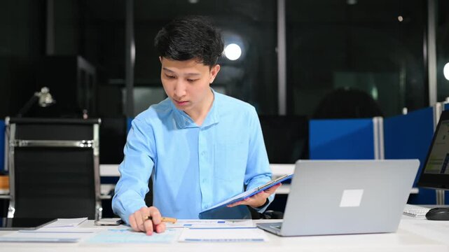 Young Asian businessman using a smartphone app for digital verification while working on a laptop late