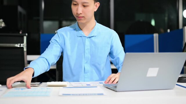 Asian businessman checking data report on a laptop and writing notes on a clipboard in the office.