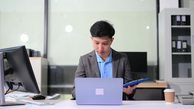 Asian businessman checking data report on a laptop and writing notes on a clipboard in the office.