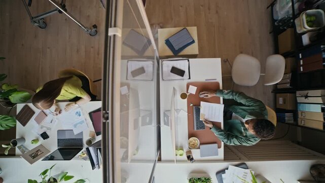 Top view of coworkers using laptops and reviewing documents at separate desks in contemporary corporate office