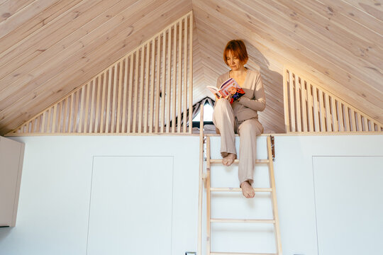 Mature Woman Reading Book In Cozy Loft Bedroom Of Modern Tiny House