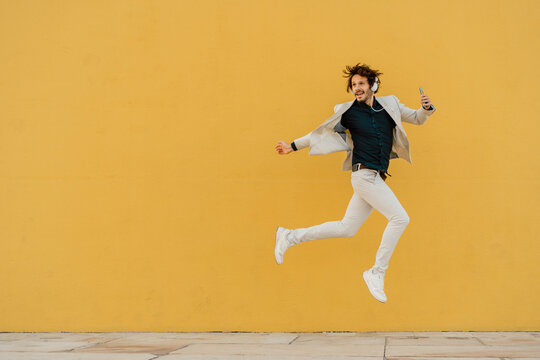 Businessman jumping in the air in front of yellow wall listening music with headphones and smartphone
