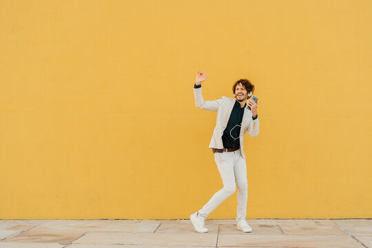 Businessman singing and dancing in front of yellow wall listening music with headphones and smartphone