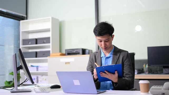 Asian businessman checking data report on a laptop and writing notes on a clipboard in the office.