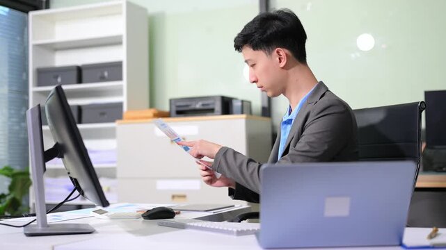 Asian businessman checking data report on a laptop and writing notes on a clipboard in the office.