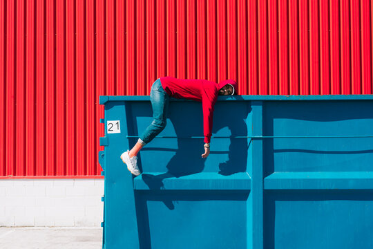 Young man wearing red hooded jacket lying on edge of container in front of red wall
