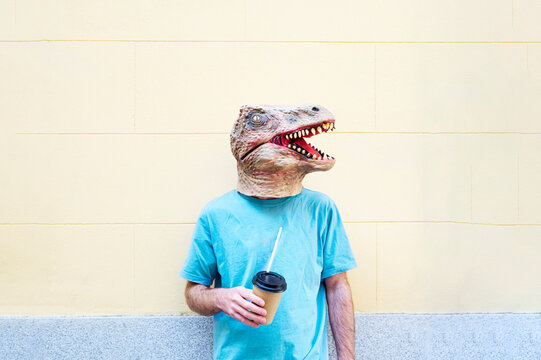 Mid adult man wearing dinosaur mask with coffee standing against wall in city