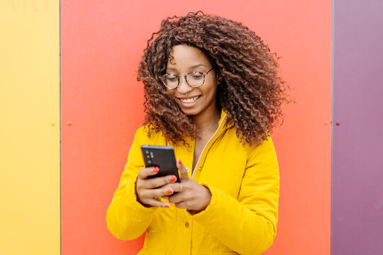 Young woman wearing jacket using mobile phone while standing against multi colored wall
