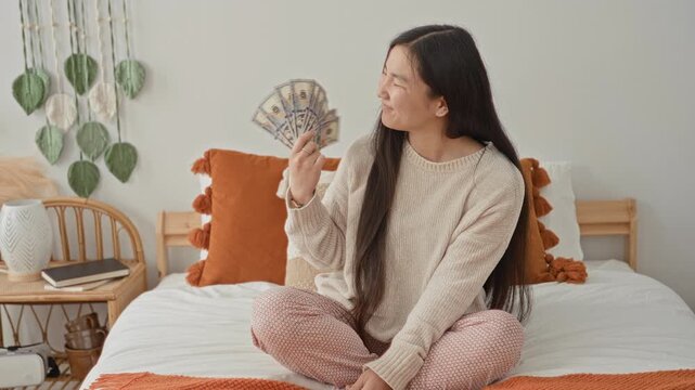 Woman fanning a stack of dollar bills on bed, sitting crosslegged, smiling while holding money in hand in a cozy bedroom with pillows and a rattan nightstand; joy abundance.