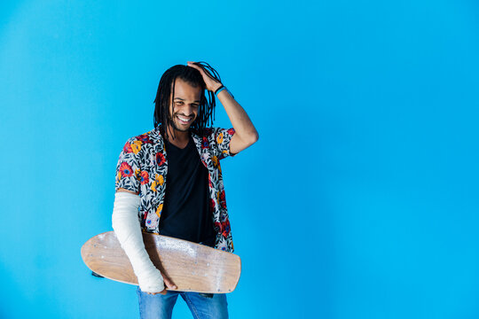 Smiling man with fractured arm holding skateboard against blue background
