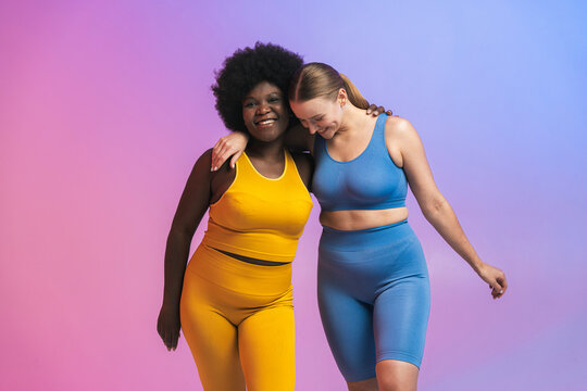 Happy young multiracial female friends standing with arms around against colored background