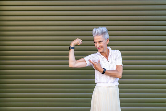 Active senior woman pointing at bicep while flexing muscles while standing against shutter