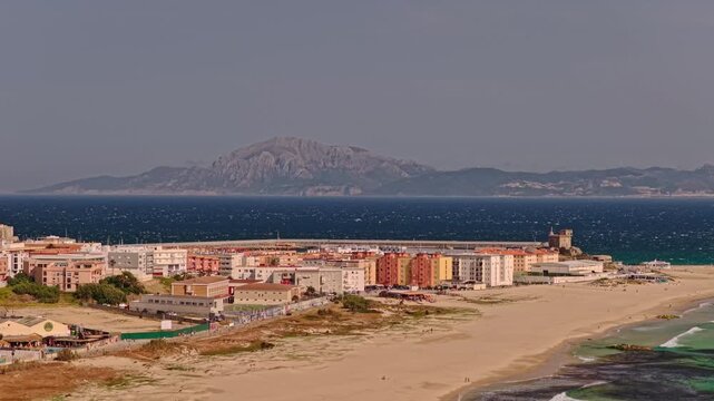 Cinematic telephoto drone orbit around Tarifa, Spain with dramatic lens compression revealing Jebel Musa in Morocco rising across the Strait of Gibraltar above the Atlantic coastline.