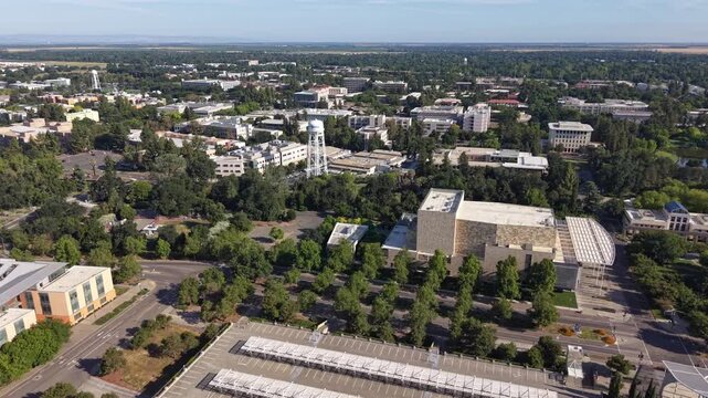 Aerial daytime drone view of the University of California, Davis, campus, showing academic buildings, parking lots with solar panels, green trees, and the city of Davis in the background